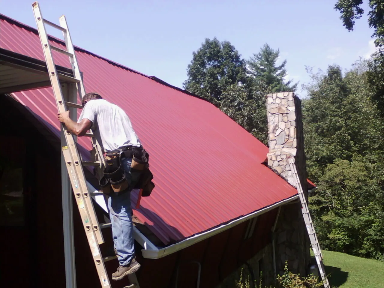 Roofer on a ladder beside a red metal roof and stone chimney — Soffit & Fascia Repair by Roofing Brothers in Ucon